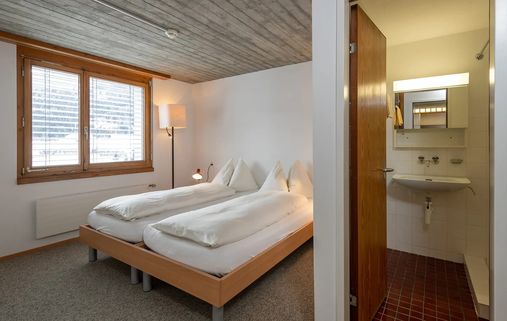 Zweibettzimmer mit Holzdecke und Fenster, daneben der Blick ins Badezimmer