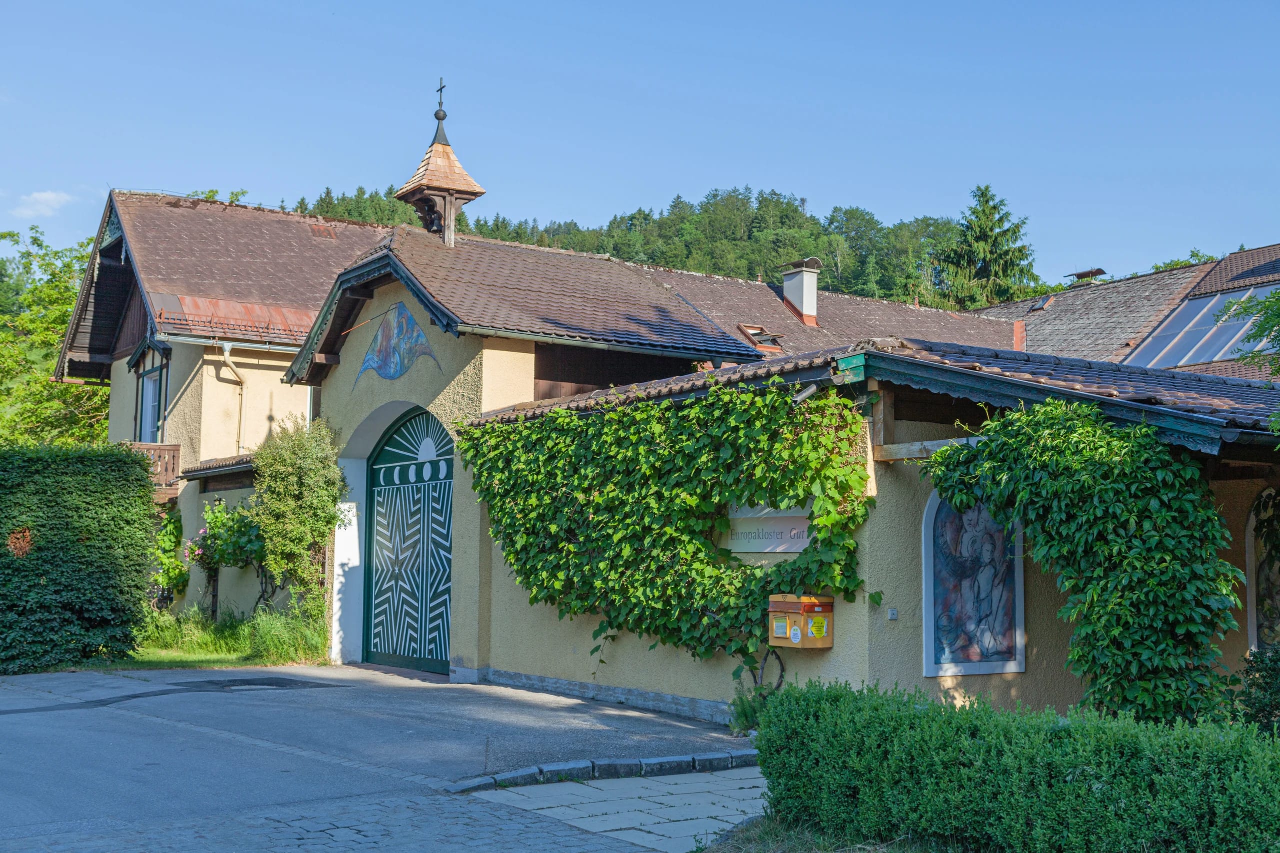 Außenansicht des gelben Klostergebäudes mit begrüntem Rundbogentor, Dachreiter mit Kreuz und bewaldetem Hang dahinter