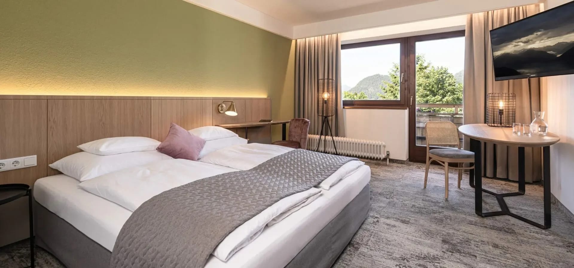 Modernes Doppelzimmer mit grauem Doppelbett, Schreibtisch und Fenster mit Bergblick