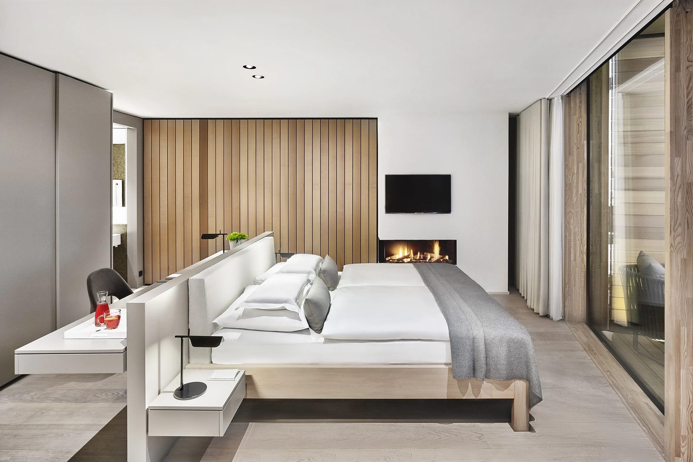Suite mit Bett vor holzverkleideter Wand und offenem Kamin, seitlich eine raumhohe Verglasung