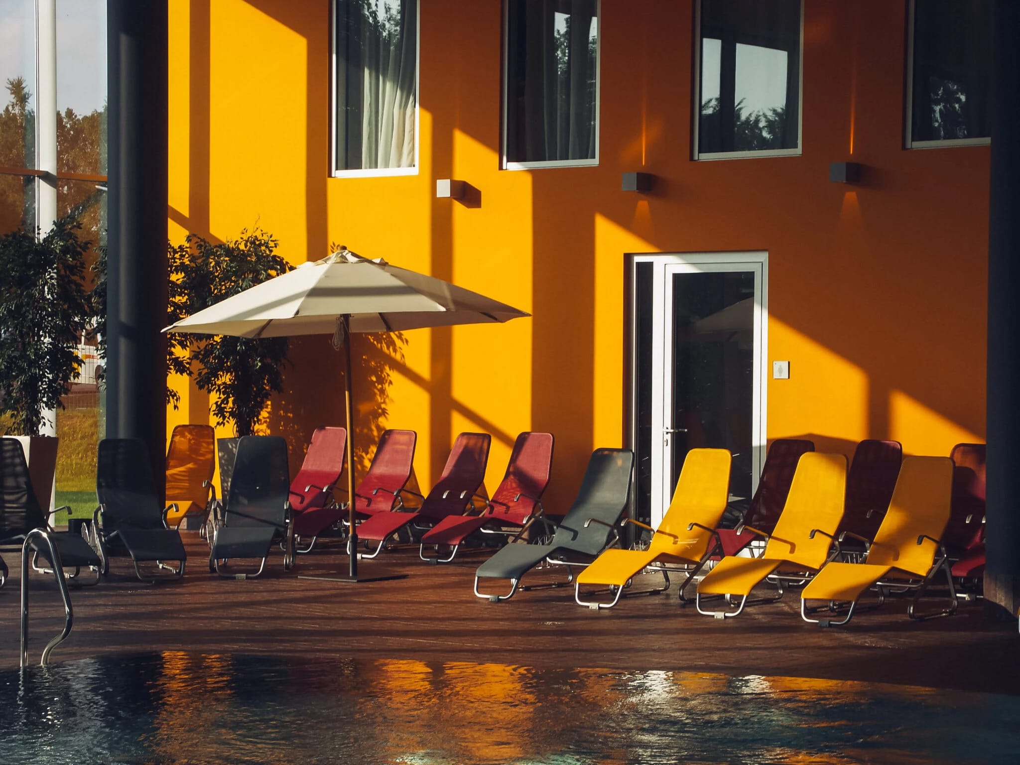 Liegen mit Sonnenschirm auf der Holzterrasse am Pool vor der orangen Hausfassade