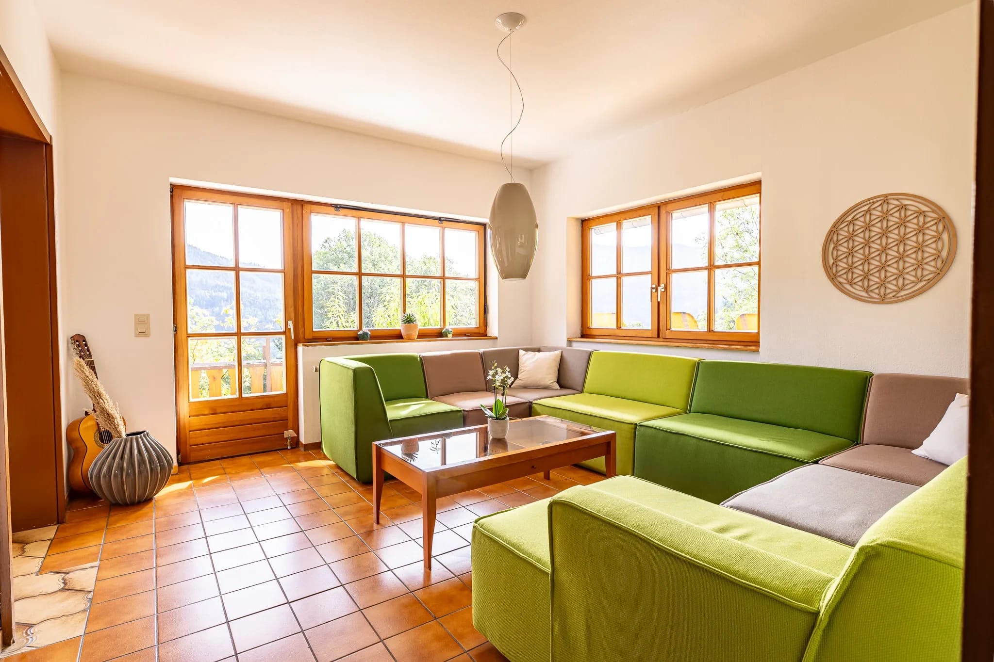 Aufenthaltsraum mit grünen Sofas, niedrigem Holztisch, Terrakottaboden und Fenstern ins Grüne