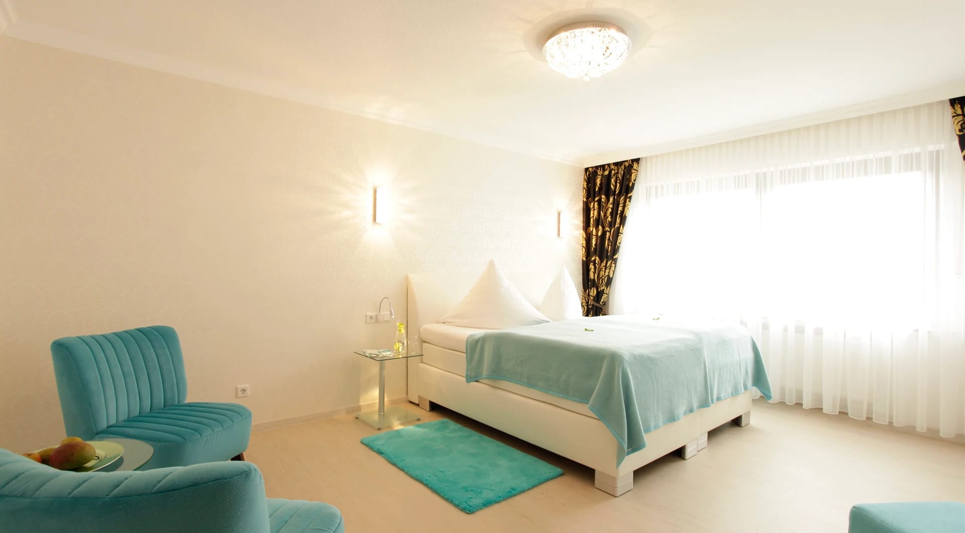Helles Zimmer mit weißem Bett, türkisfarbener Tagesdecke und türkisem Sofa