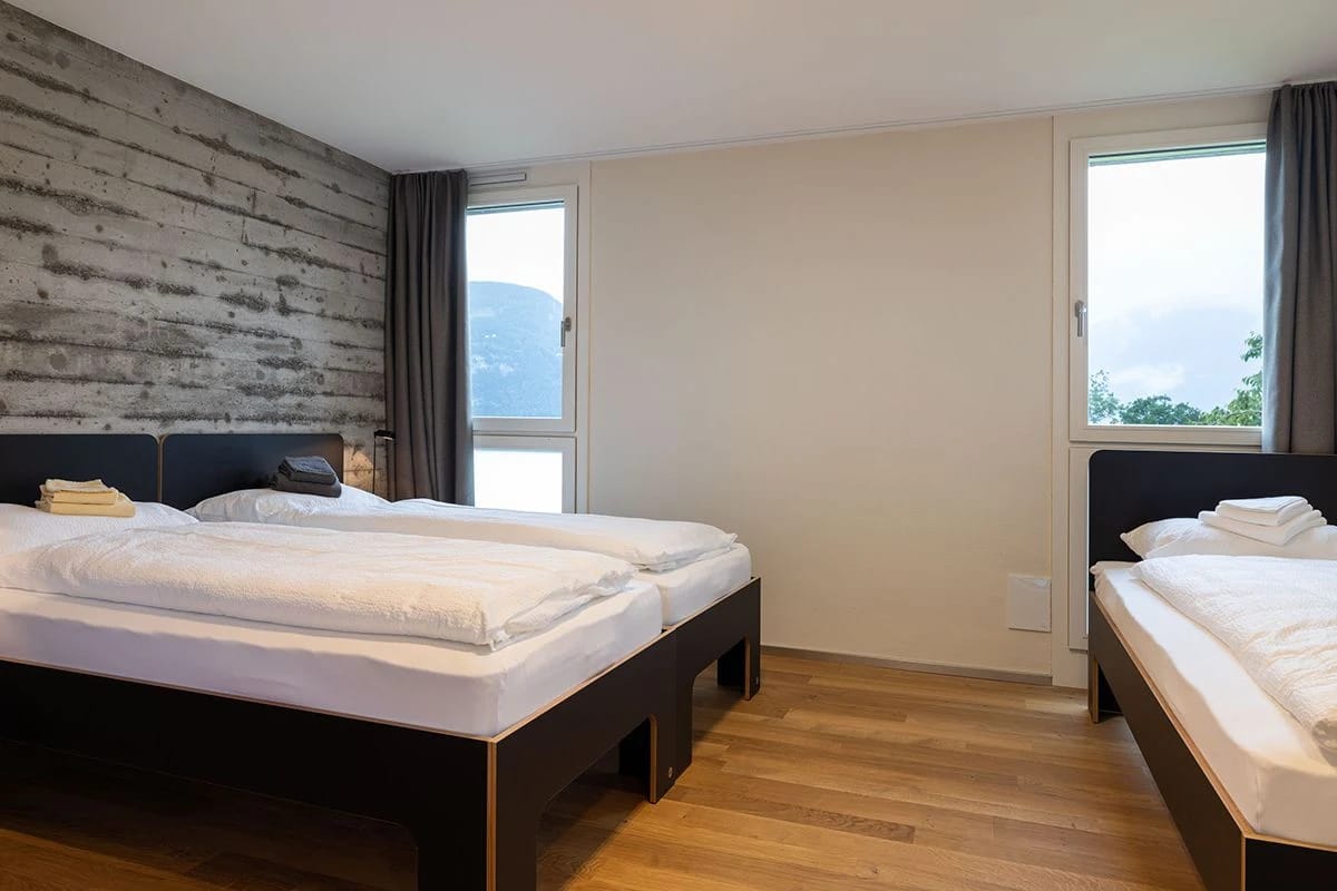 Familienzimmer mit Doppelbett und zusätzlichem Einzelbett, Sichtbetonwand und zwei Fenstern