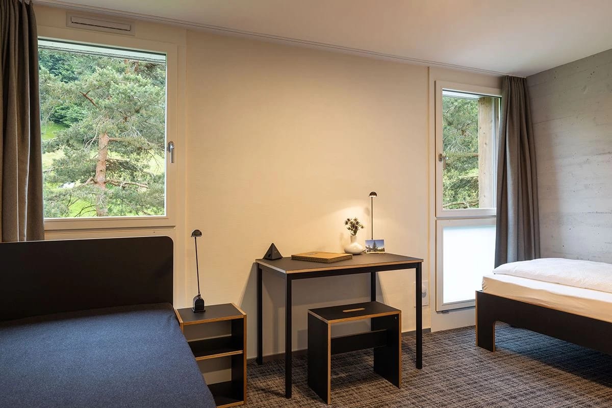 Zimmer mit Schreibtisch zwischen zwei Fenstern mit Blick in den Wald und Einzelbett