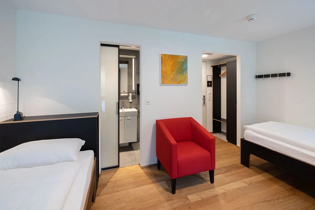 Zweibettzimmer mit rotem Sessel, Holzboden und offener Tür zum Badezimmer