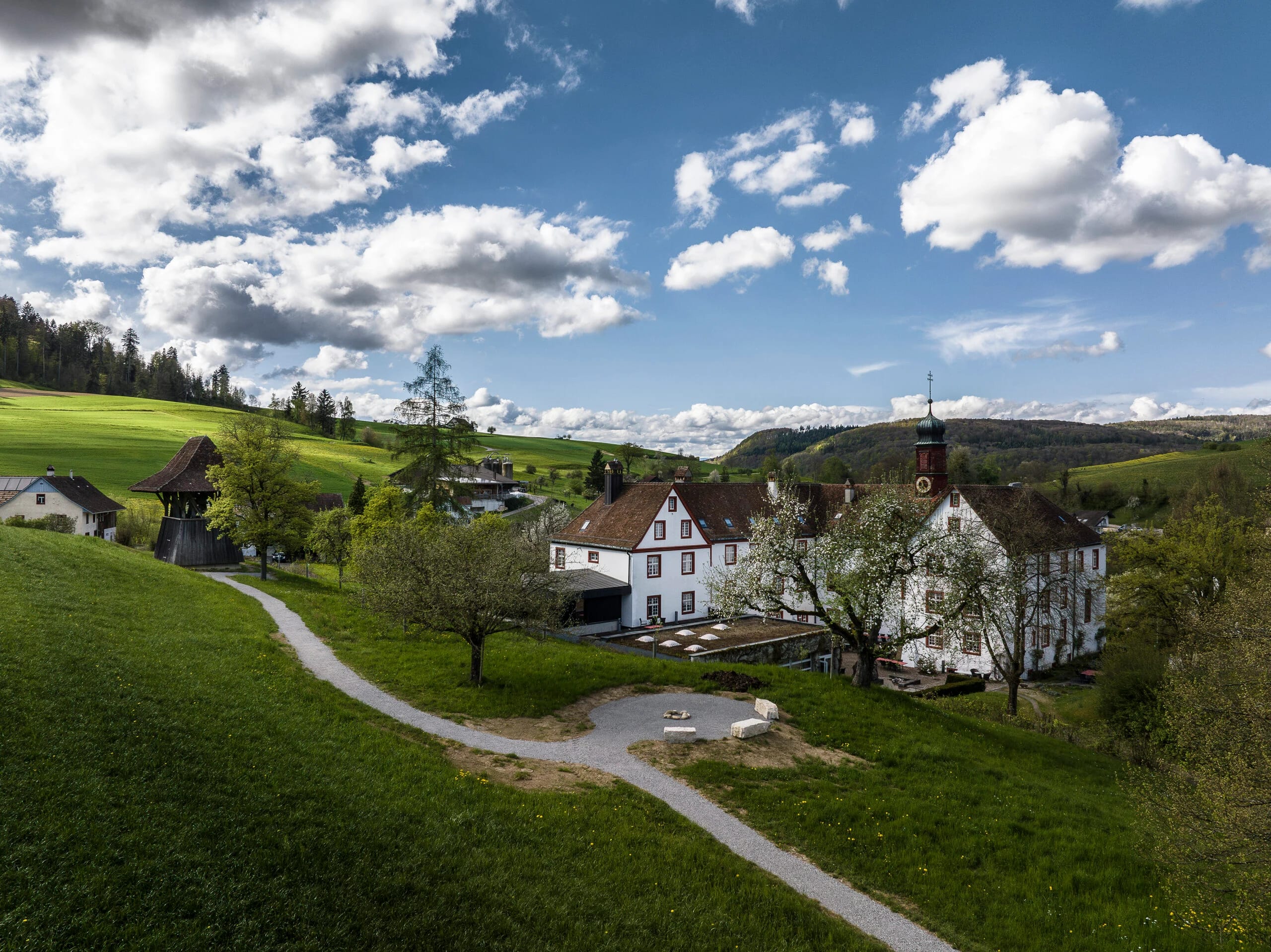 Blick über Wiesen und geschwungene Wege auf die Klosteranlage in hügeliger Landschaft