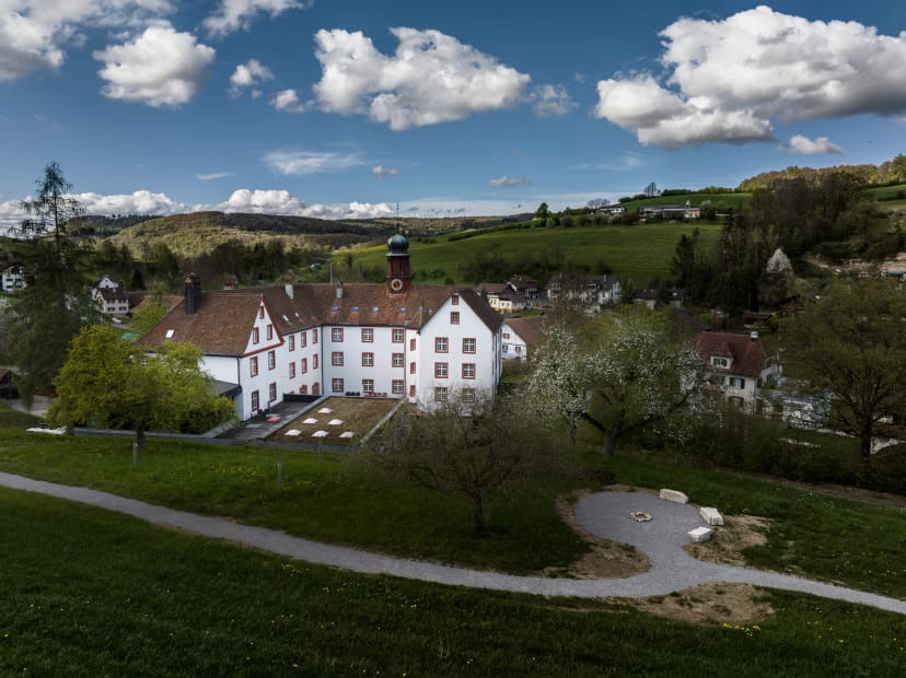 Weiss verputzte Klosteranlage mit Zwiebelturm, davor Wiese und blühender Baum unter blauem Himmel