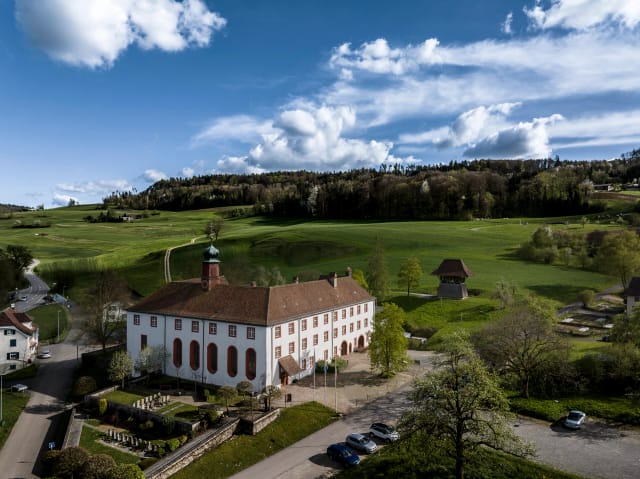 Luftaufnahme der Klosteranlage mit Kirchenflügel, eingebettet in grüne Felder und Hügel