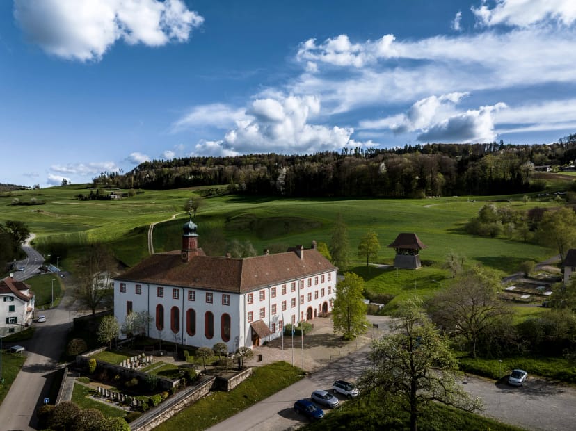 Luftaufnahme der Klosteranlage mit Kirchenflügel, eingebettet in grüne Felder und Hügel