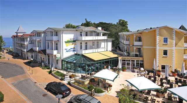 Hotelensemble mit weißen Häusern und gelbem Gebäude, davor Terrasse mit Sonnenschirmen und verglaster Wintergarten, links die Ostsee