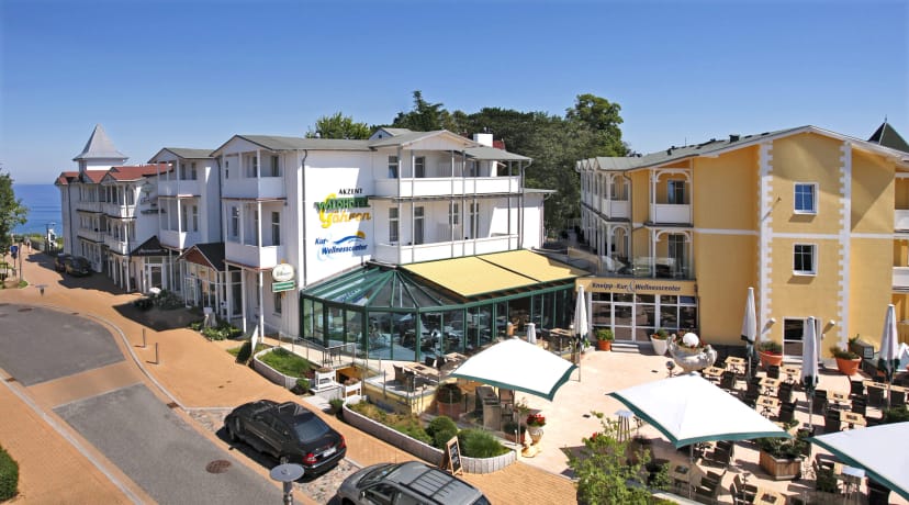 Hotelensemble mit weißen Häusern und gelbem Gebäude, davor Terrasse mit Sonnenschirmen und verglaster Wintergarten, links die Ostsee