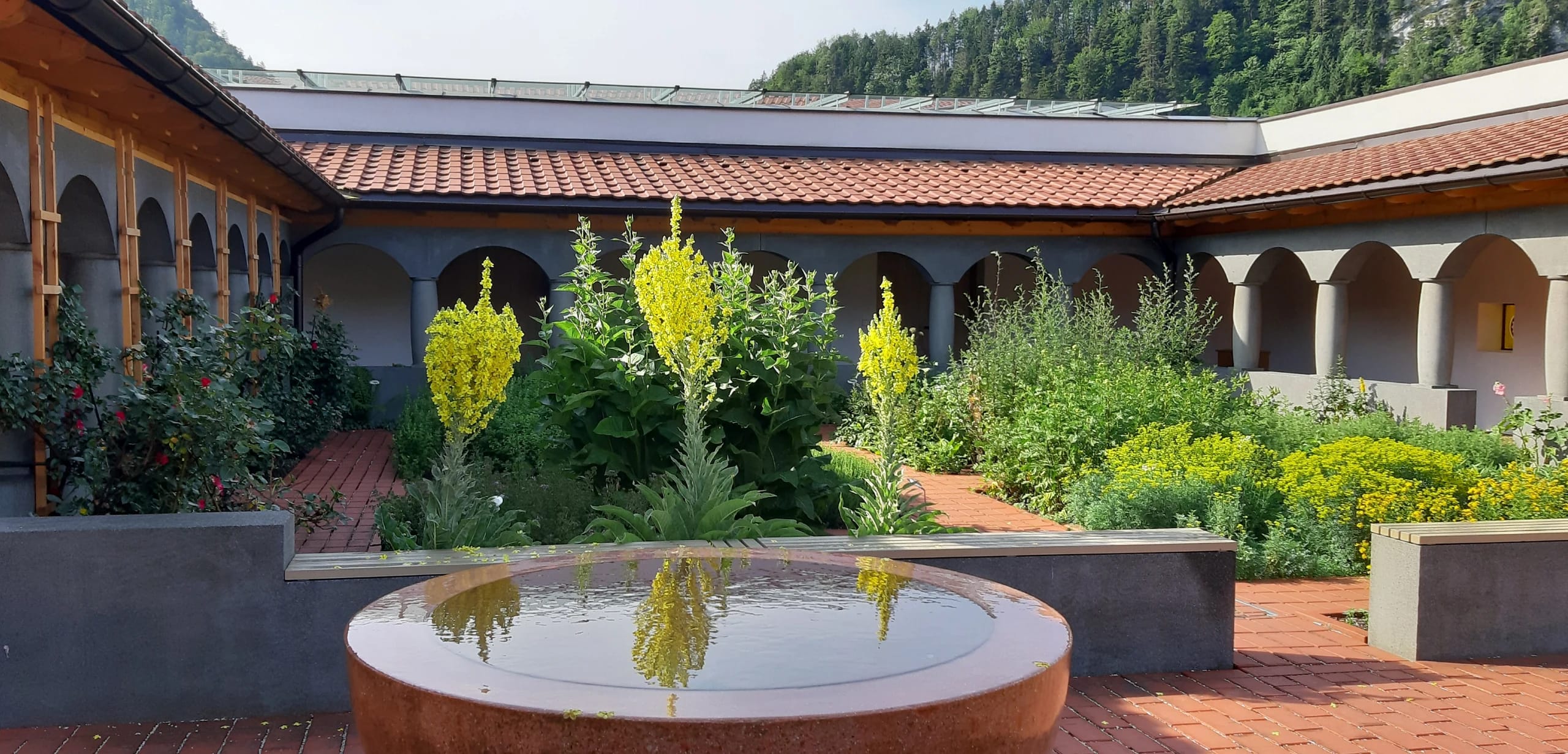 Innenhof mit Kräuterbeeten, rundem Wasserbecken und Arkadengang vor bewaldetem Berghang