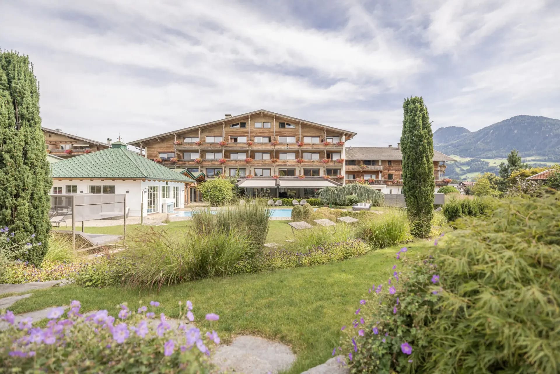 Blick auf das Hotel Pirchnerhof vom Garten aus mit Blumenbeeten, Außenpool und Bergkulisse