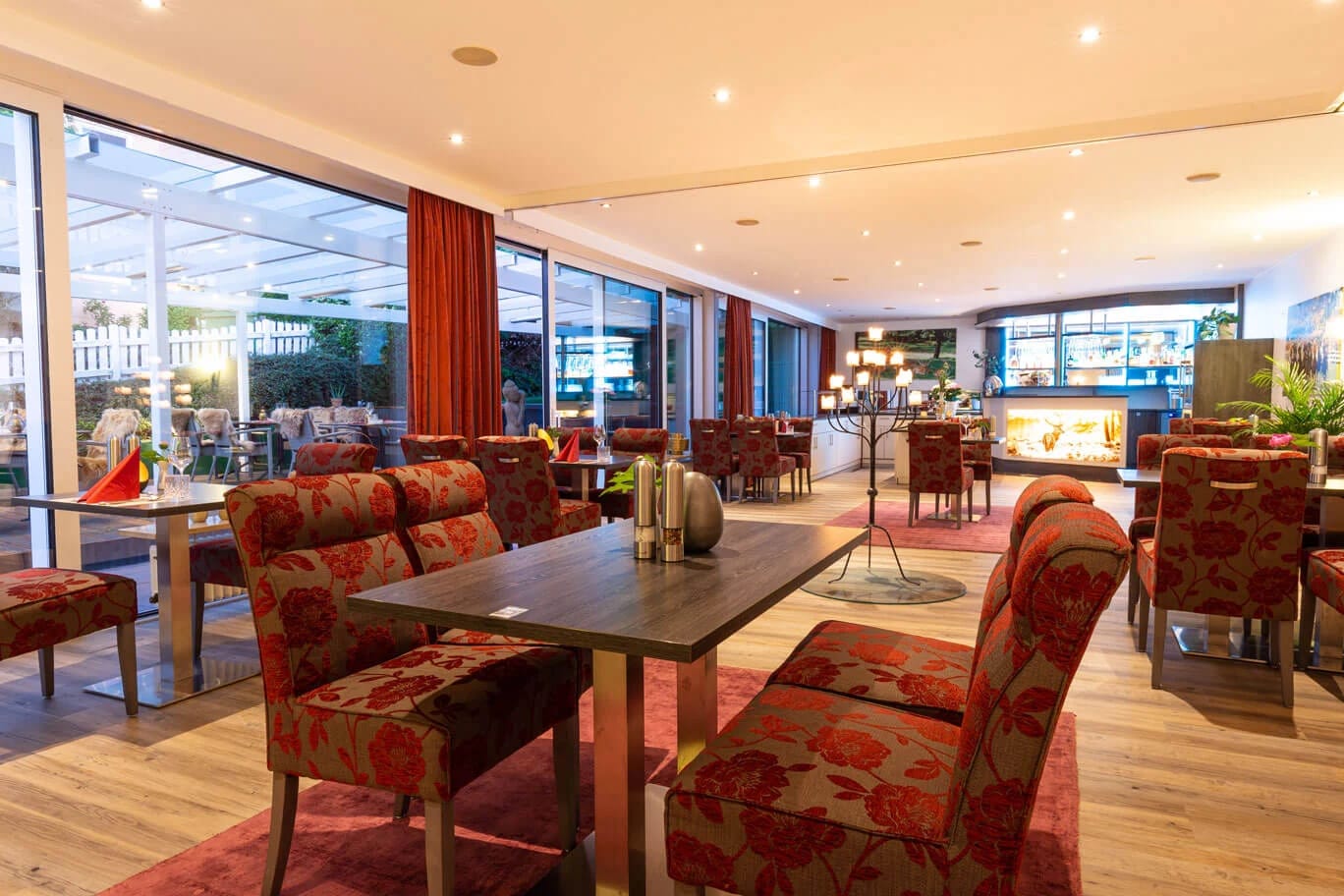 Restaurantbereich mit rot gemusterten Polstersesseln, Fensterfront und Blick auf die beleuchtete Terrasse