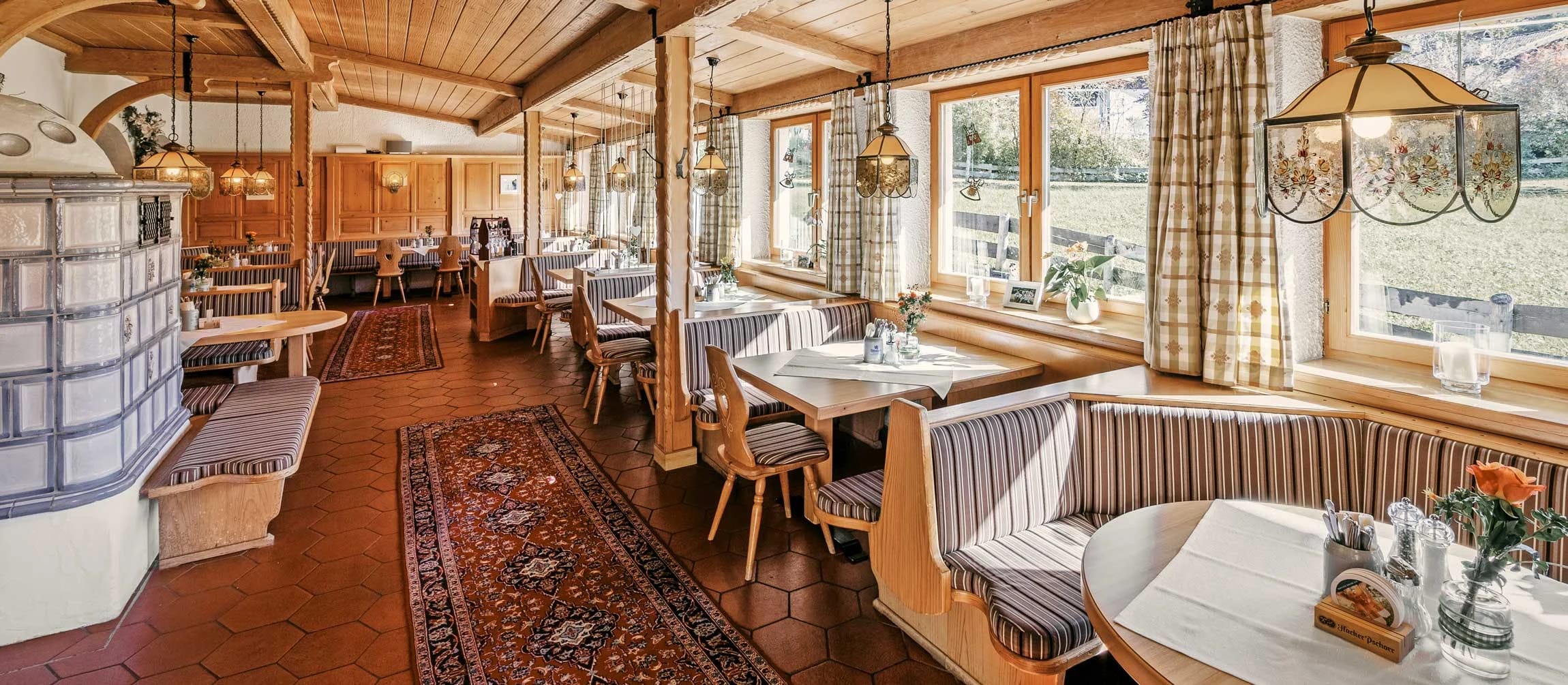 Gaststube mit Holzvertäfelung, Kachelofen und eingedeckten Tischen an den Fenstern