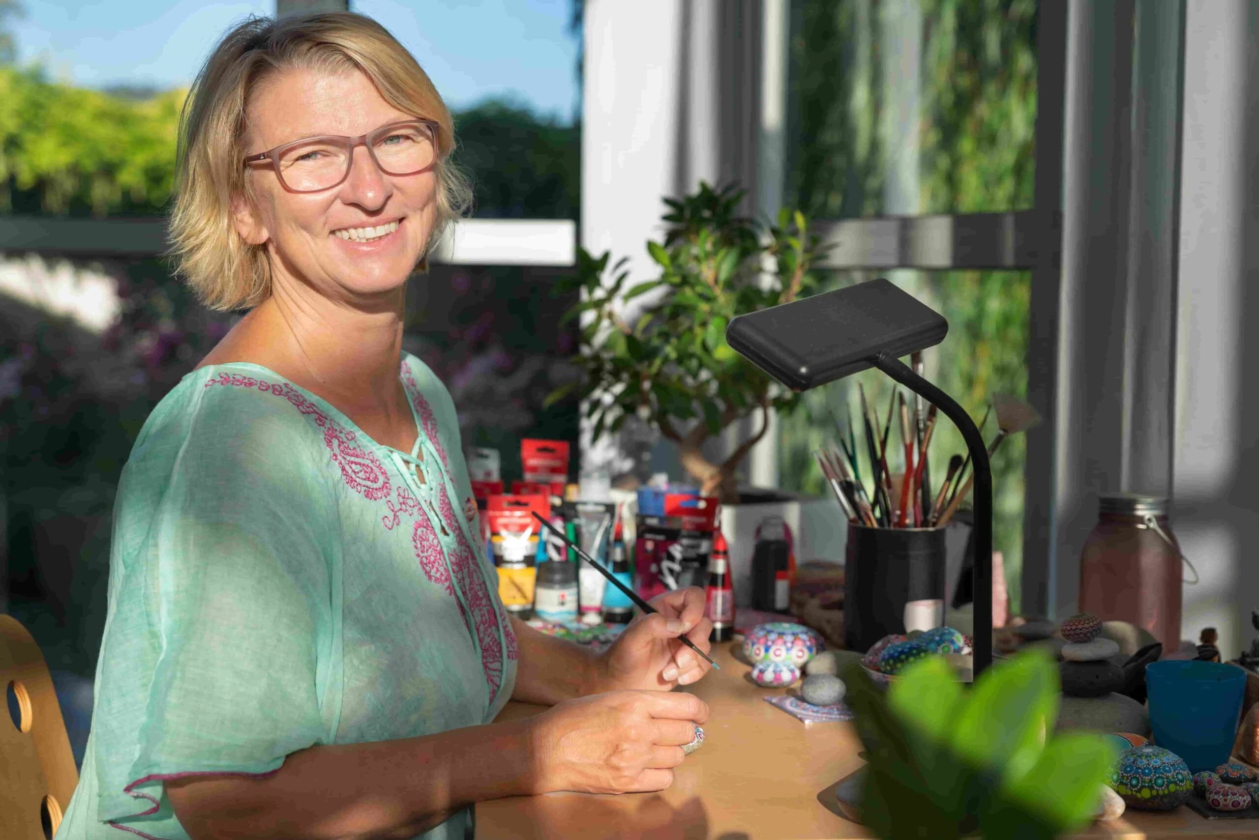 Sabine Wiemann bemalt lächelnd einen Mandalastein an einem Tisch