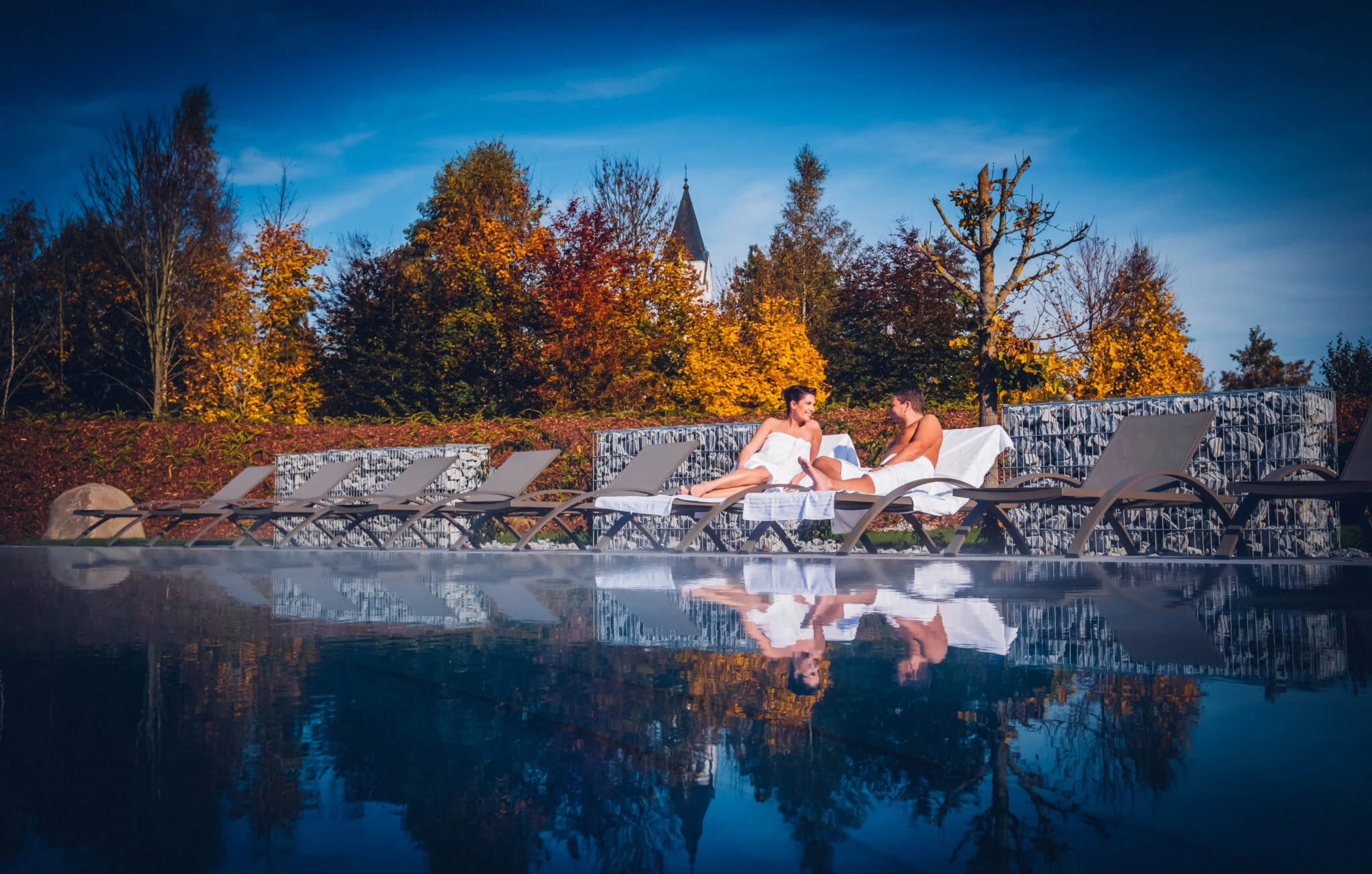 Sonnenliegen am Außenpool vor herbstlich gefärbten Bäumen, die sich im Wasser spiegeln