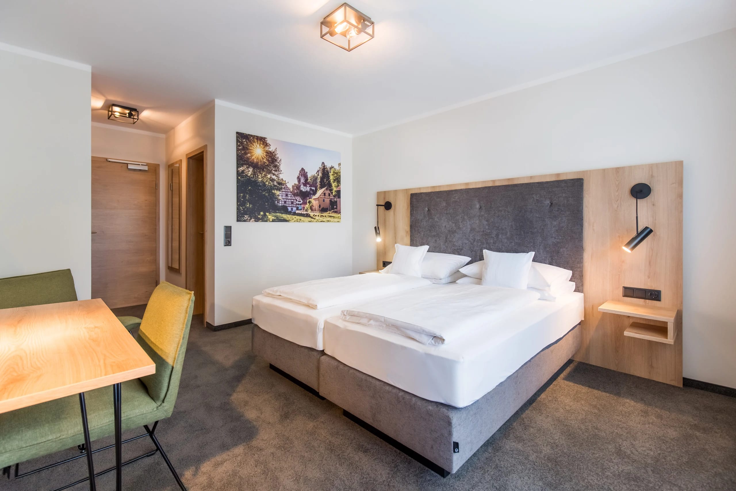 Doppelzimmer mit Bett, Holzelementen, Tisch mit zwei Stühlen und gerahmtem Wandbild