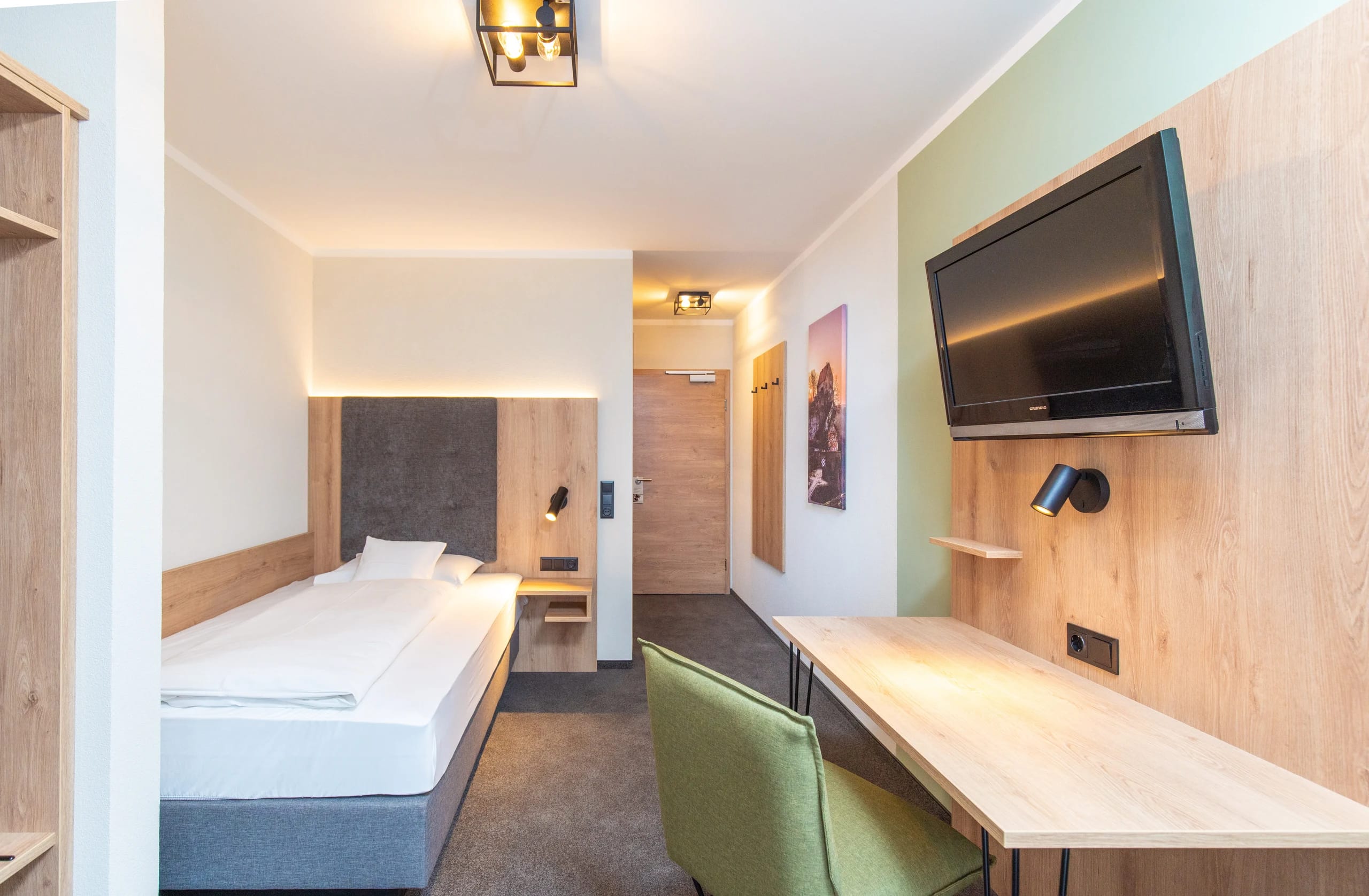 Einzelzimmer mit Bett, langem Schreibtisch, grünem Stuhl und Wandfernseher
