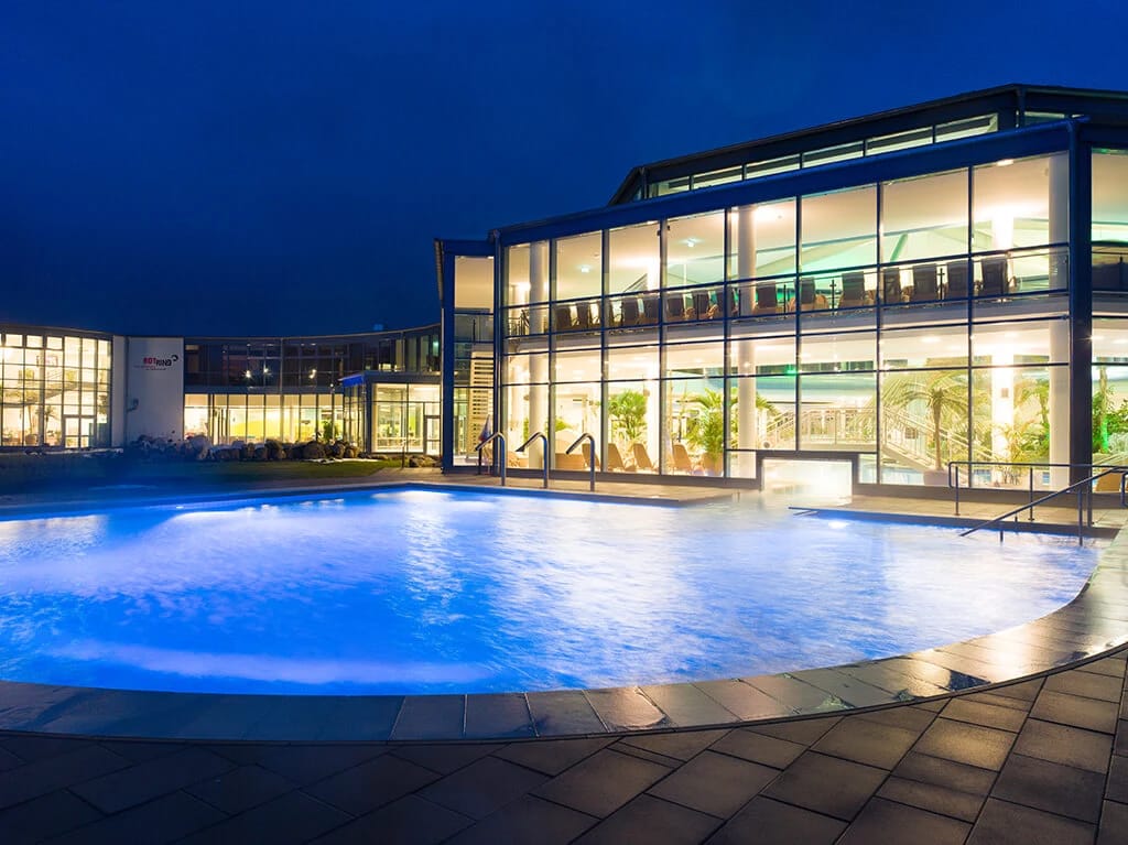 Beleuchteter Außenpool bei Nacht vor der Glasfassade der Therme