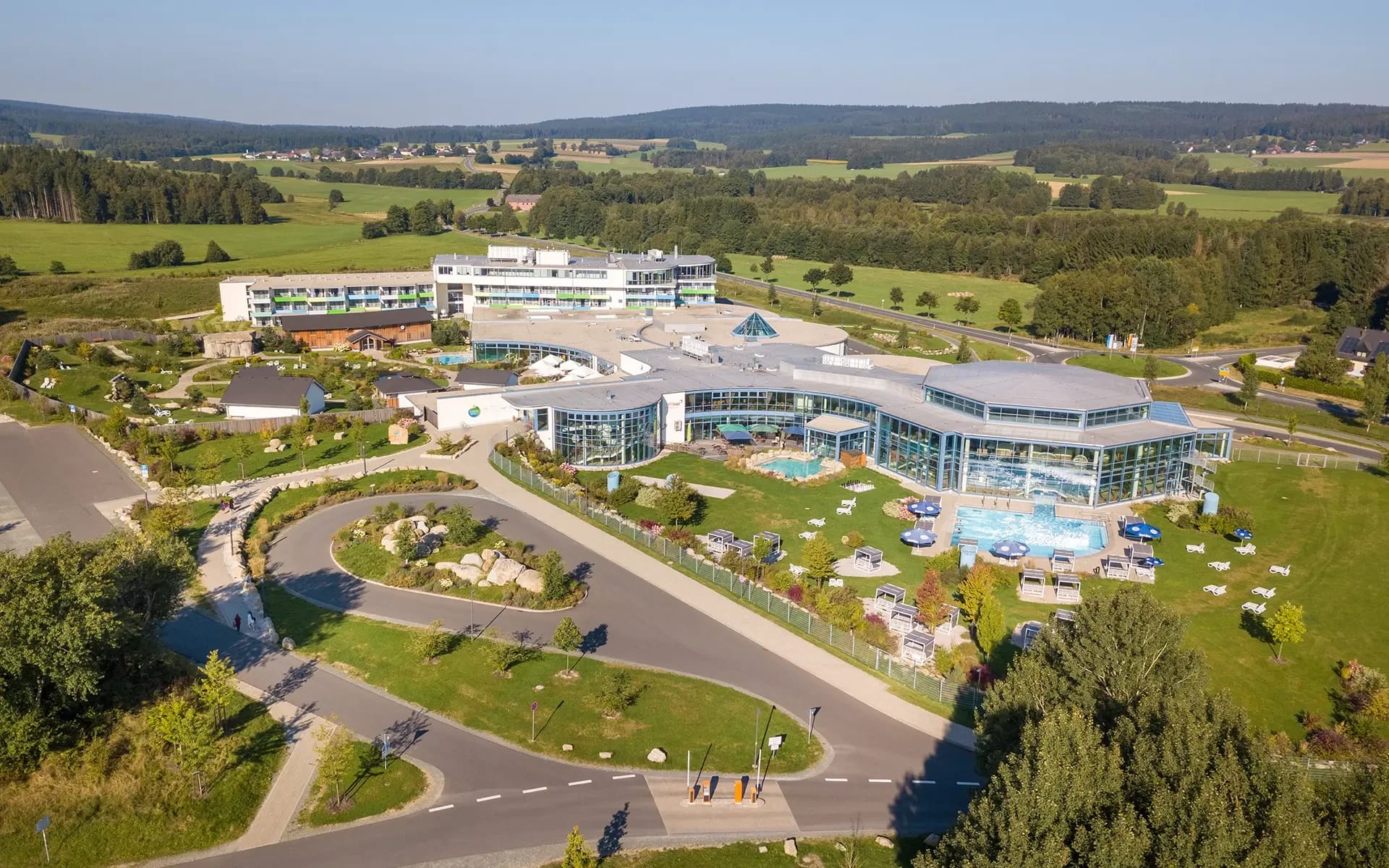 Luftaufnahme der gesamten Anlage mit langgestrecktem Hotelgebäude, Therme und Außenpool