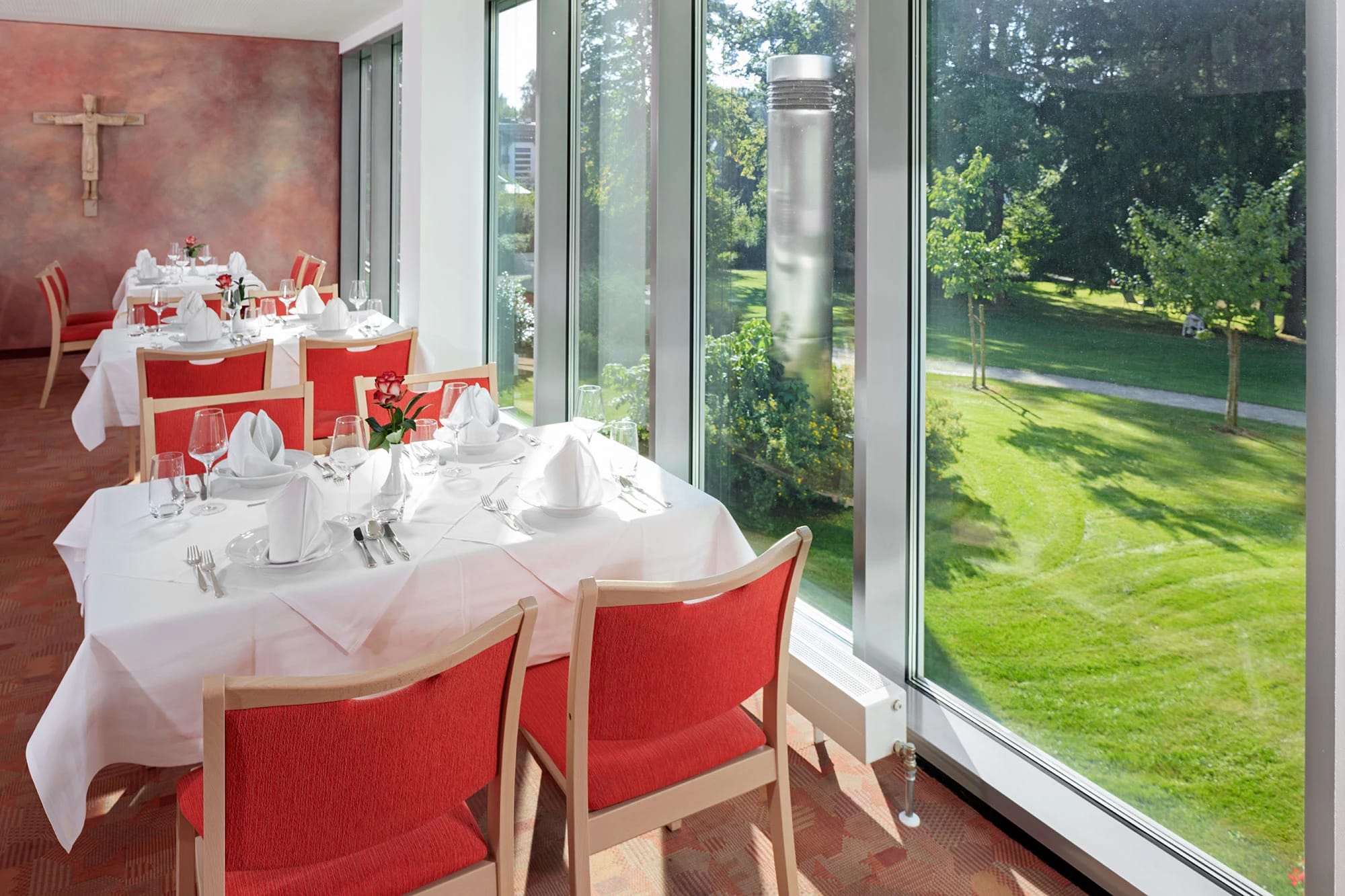Speisesaal mit eingedeckten Tischen und roten Stühlen an der Fensterfront zum Park