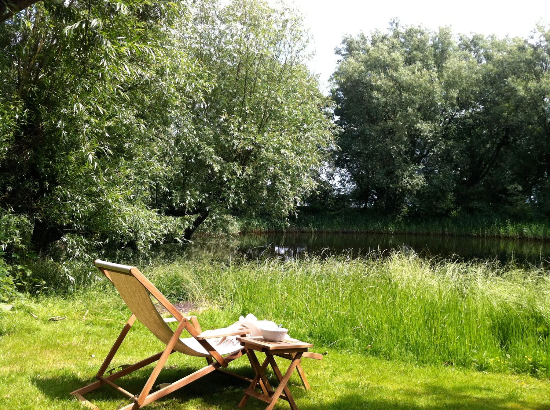 Liegestuhl mit Buch auf der Wiese am Teich, umgeben von Weiden