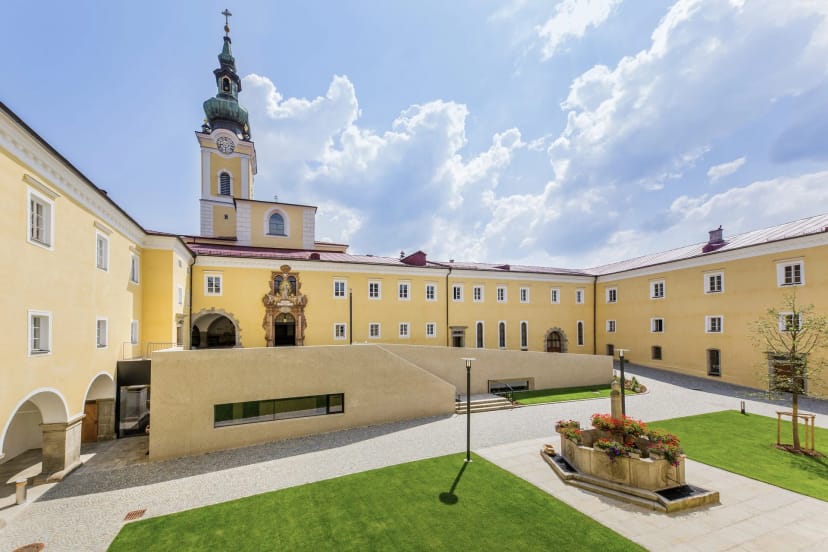 Innenhof des Stifts mit gelben Klostergebäuden, Kirchturm und Rasenflächen unter blauem Himmel