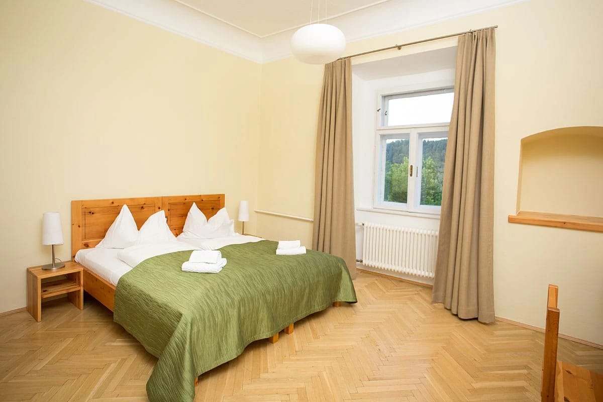 Helles Doppelzimmer mit grüner Tagesdecke, Fischgrätparkett und beigen Vorhängen