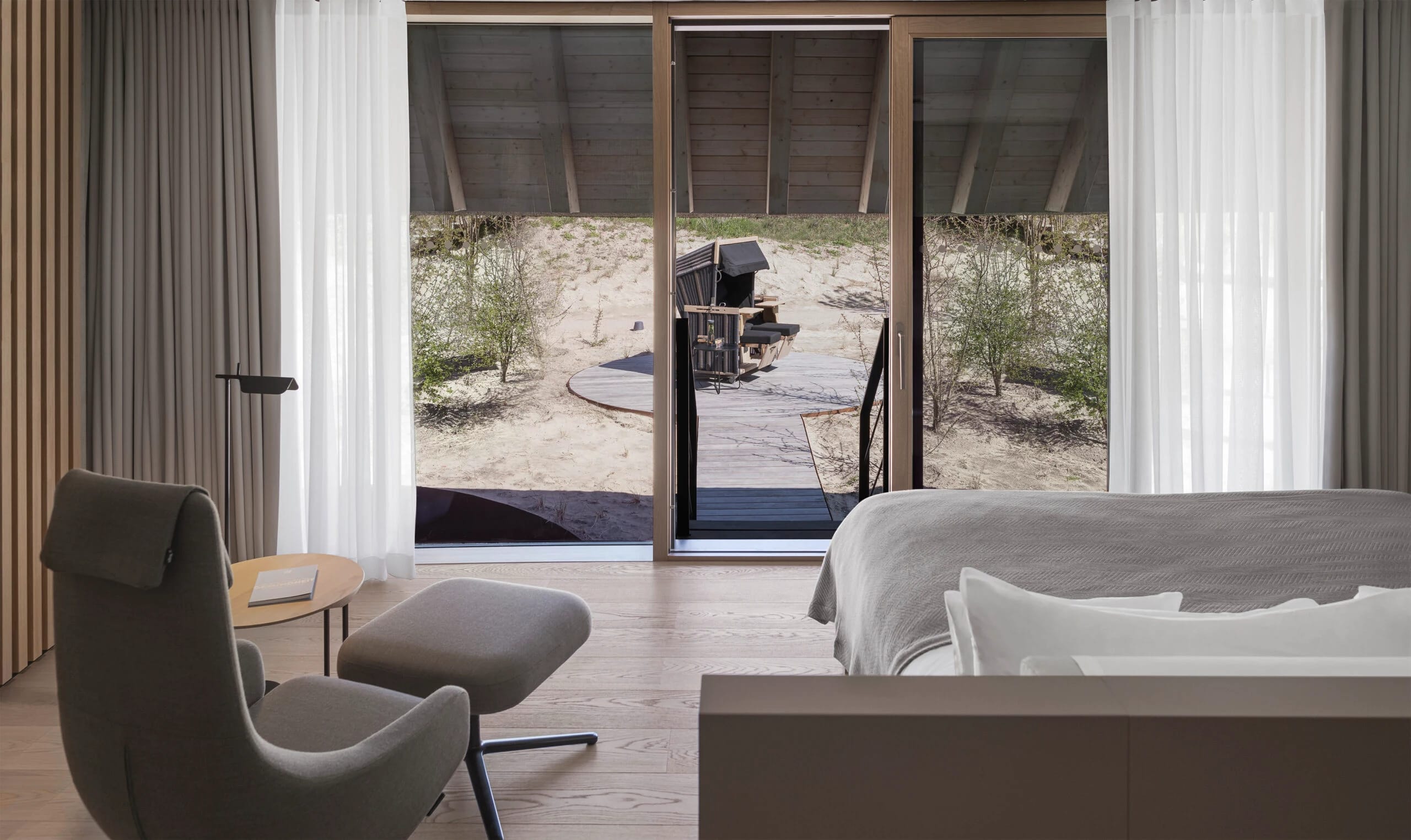 Zimmer mit Sessel und Hocker, Bett und bodentiefer Fensterfront zur Terrasse in den Dünen