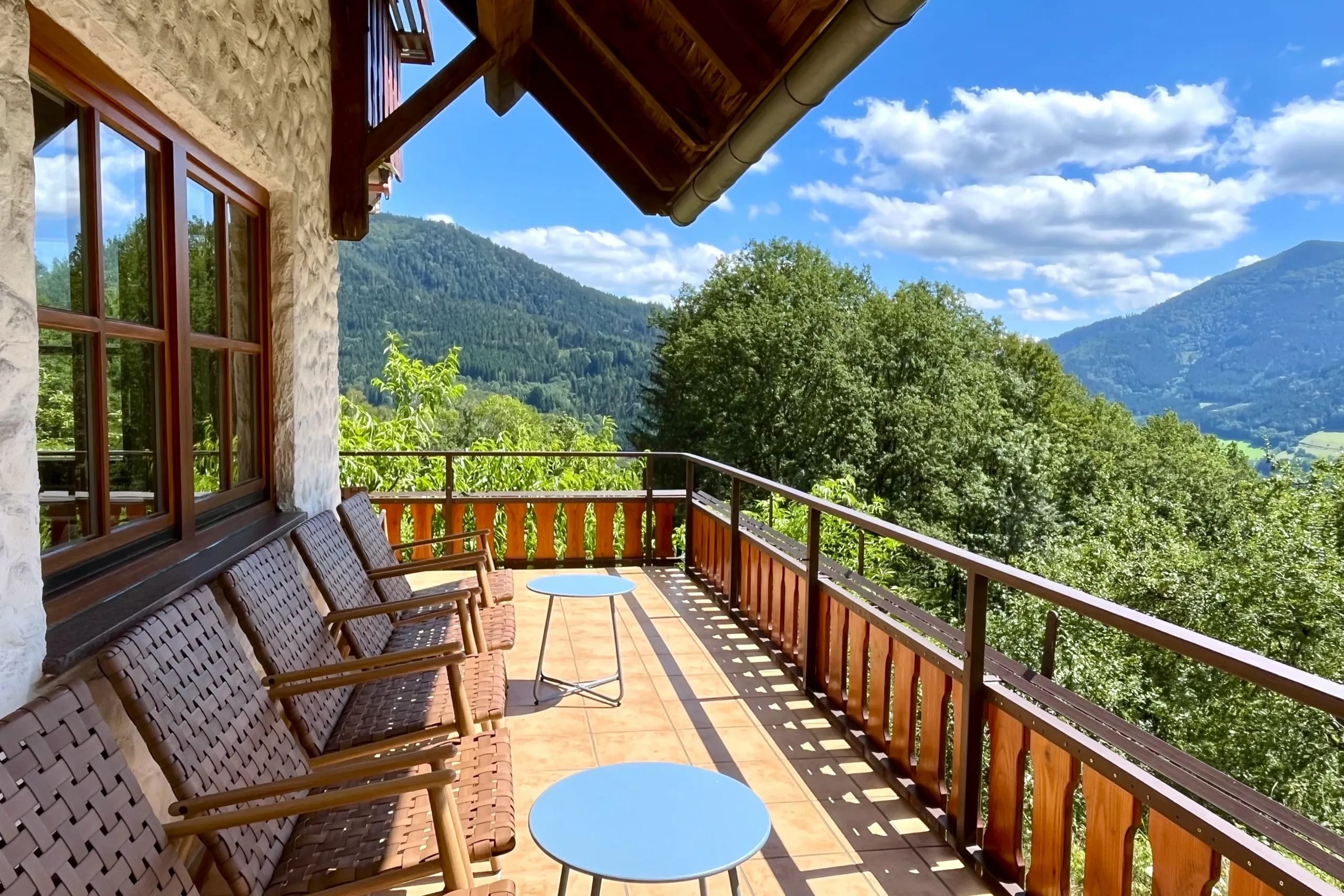 Sonniger Balkon mit Korbsesseln und runden Tischen, Holzgeländer und Blick auf die bewaldeten Berge