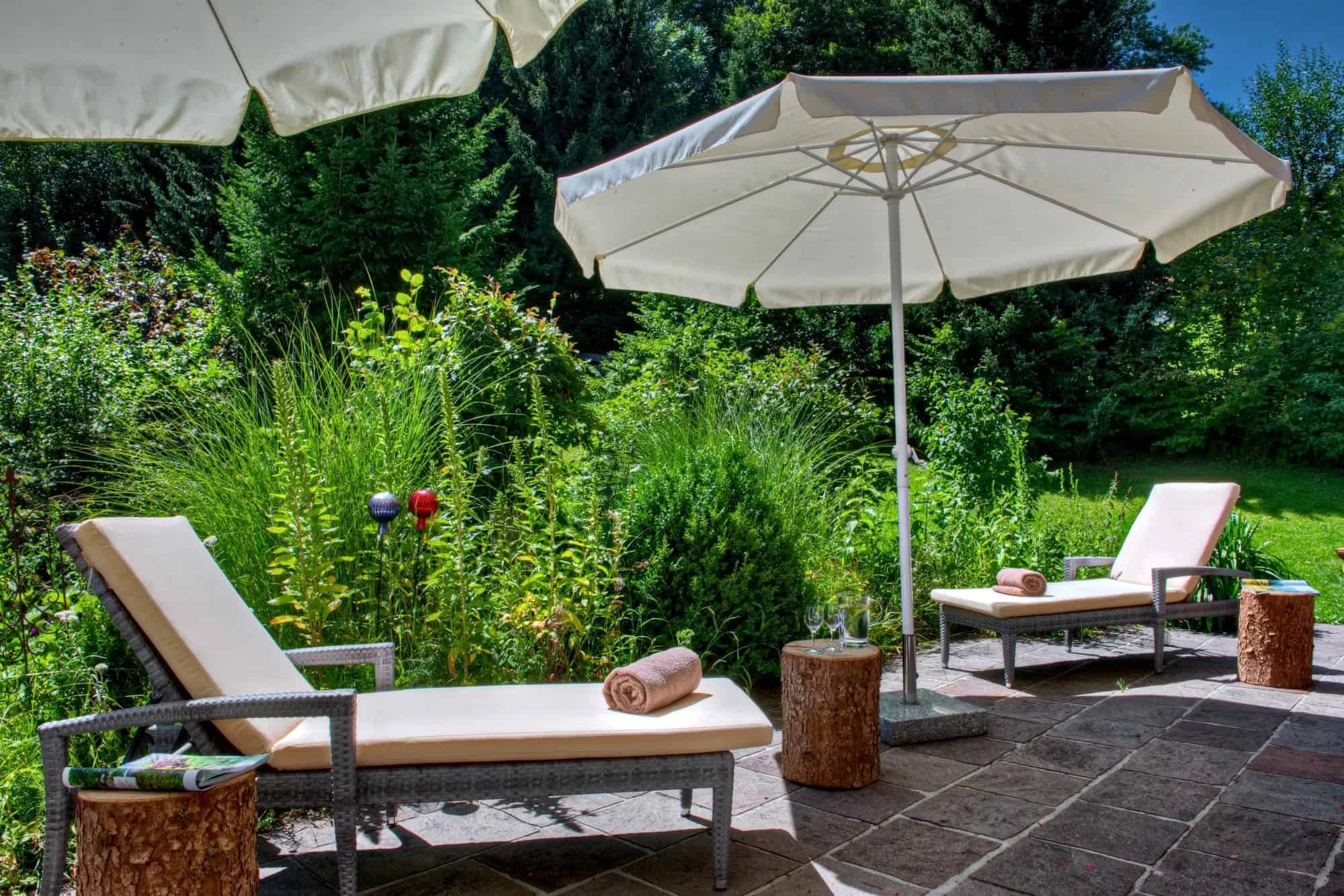 Zwei Sonnenliegen mit Sonnenschirmen auf der Gartenterrasse zwischen Sträuchern