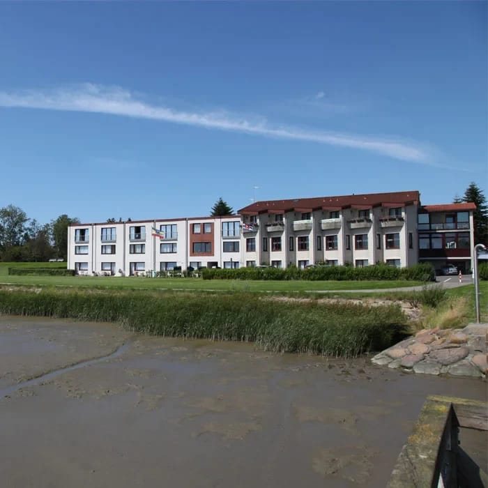 Außenansicht des langgestreckten Hotelgebäudes mit rotem Dach hinter dem Eiderdeich unter blauem Himmel