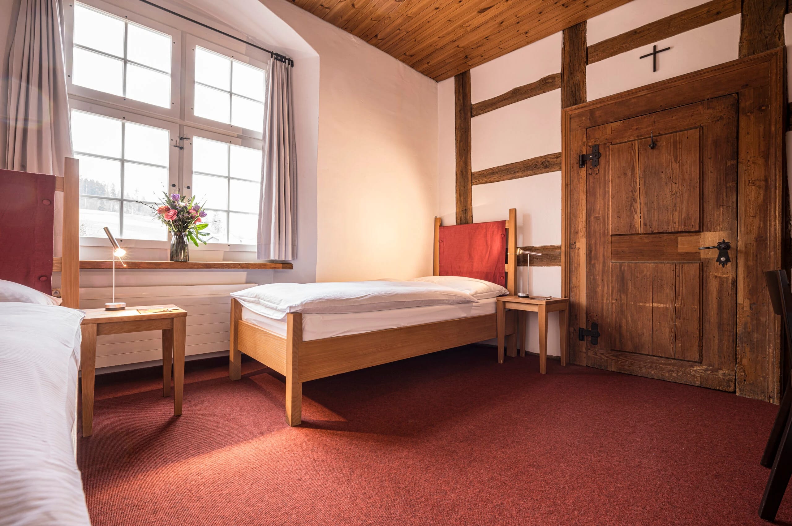 Zimmer mit rotem Teppich, Fachwerkwand und alter Holztür, Bett, Stuhl und Blumenstrauss am Fenster