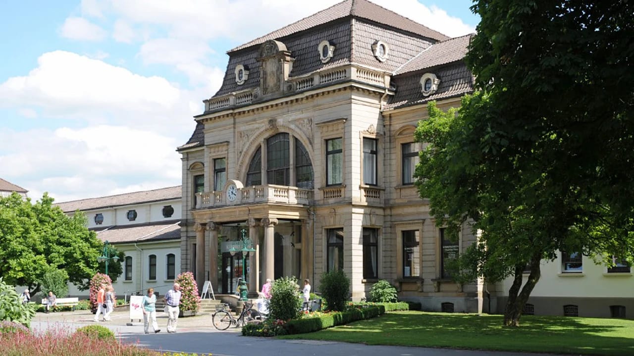 Historisches Kurhausgebäude mit Säulenvorbau, umgeben von Bäumen, davor Spaziergänger