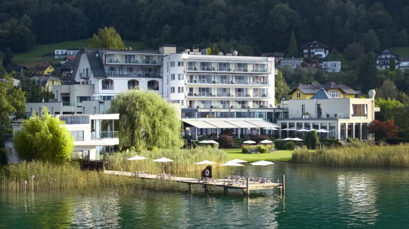 Blick vom Wörthersee auf das weiße Haupthaus mit Balkonen, davor Liegewiese, Sonnenschirme und ein Badesteg