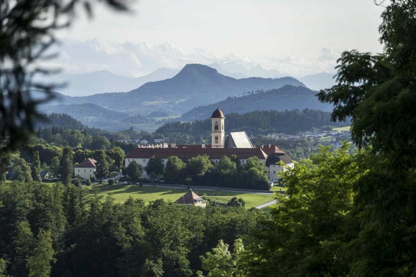 Blick über Wiesen und Wald auf das Stift mit Kirchturm, dahinter bewaldete Hügel und Berge