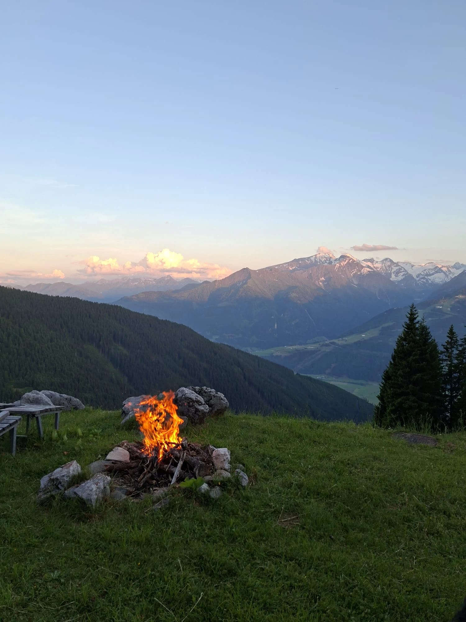 Lagerfeuer auf einer Bergwiese bei Sonnenuntergang mit Alpenpanorama