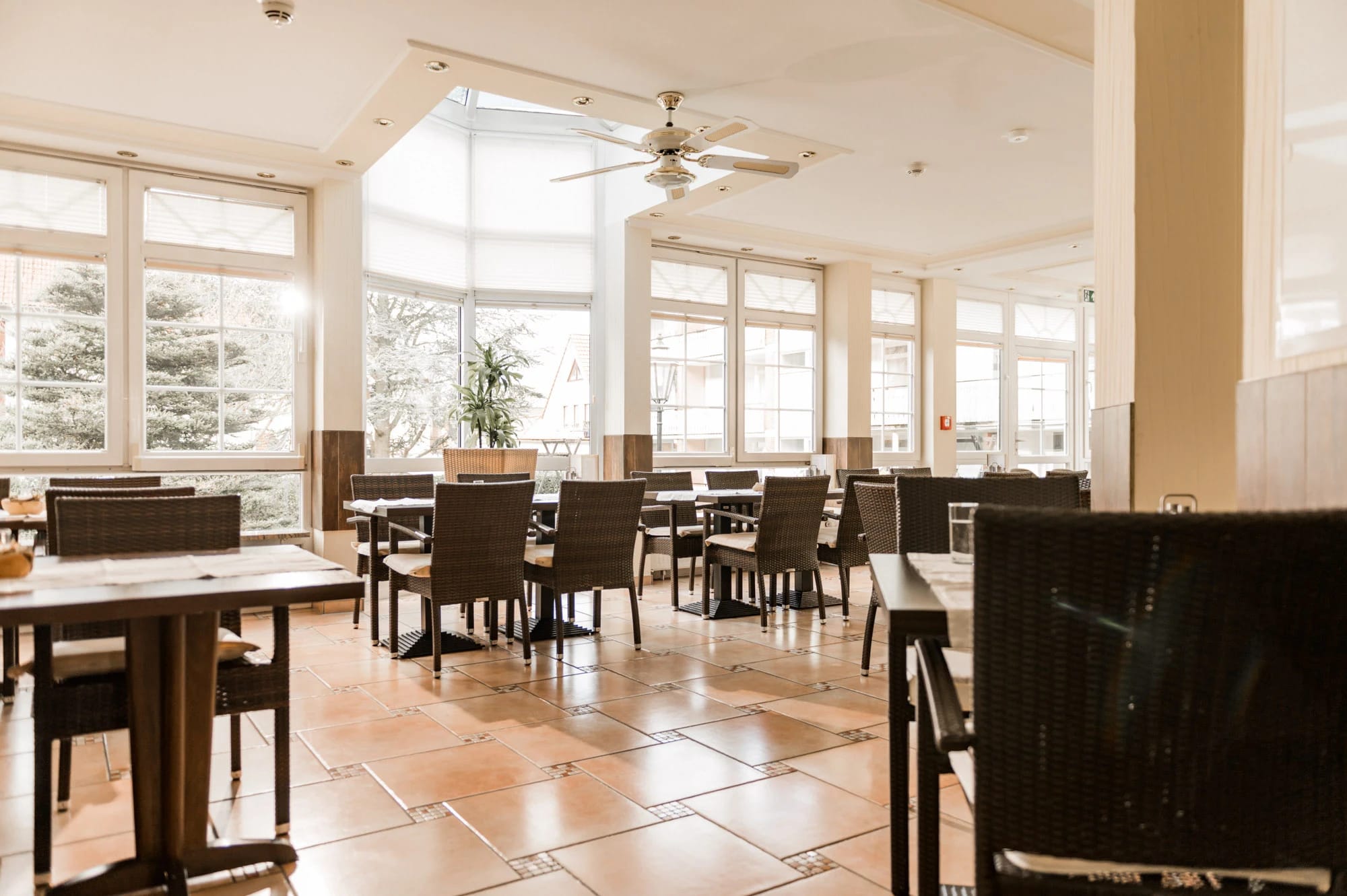 Wintergarten-Restaurant mit Korbstühlen, Fliesenboden und großen Fenstern ins Grüne