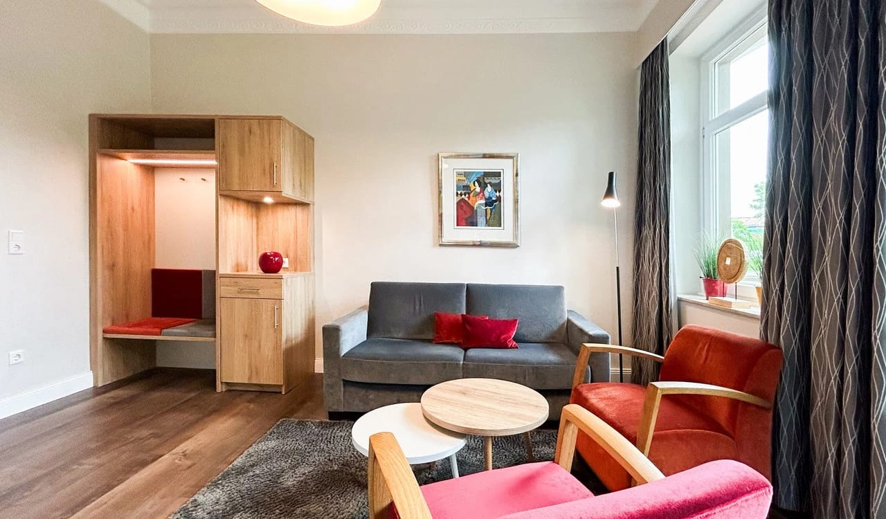 Wohnbereich mit grauem Sofa, roten Sesseln, rundem Couchtisch und heller Holzgarderobe