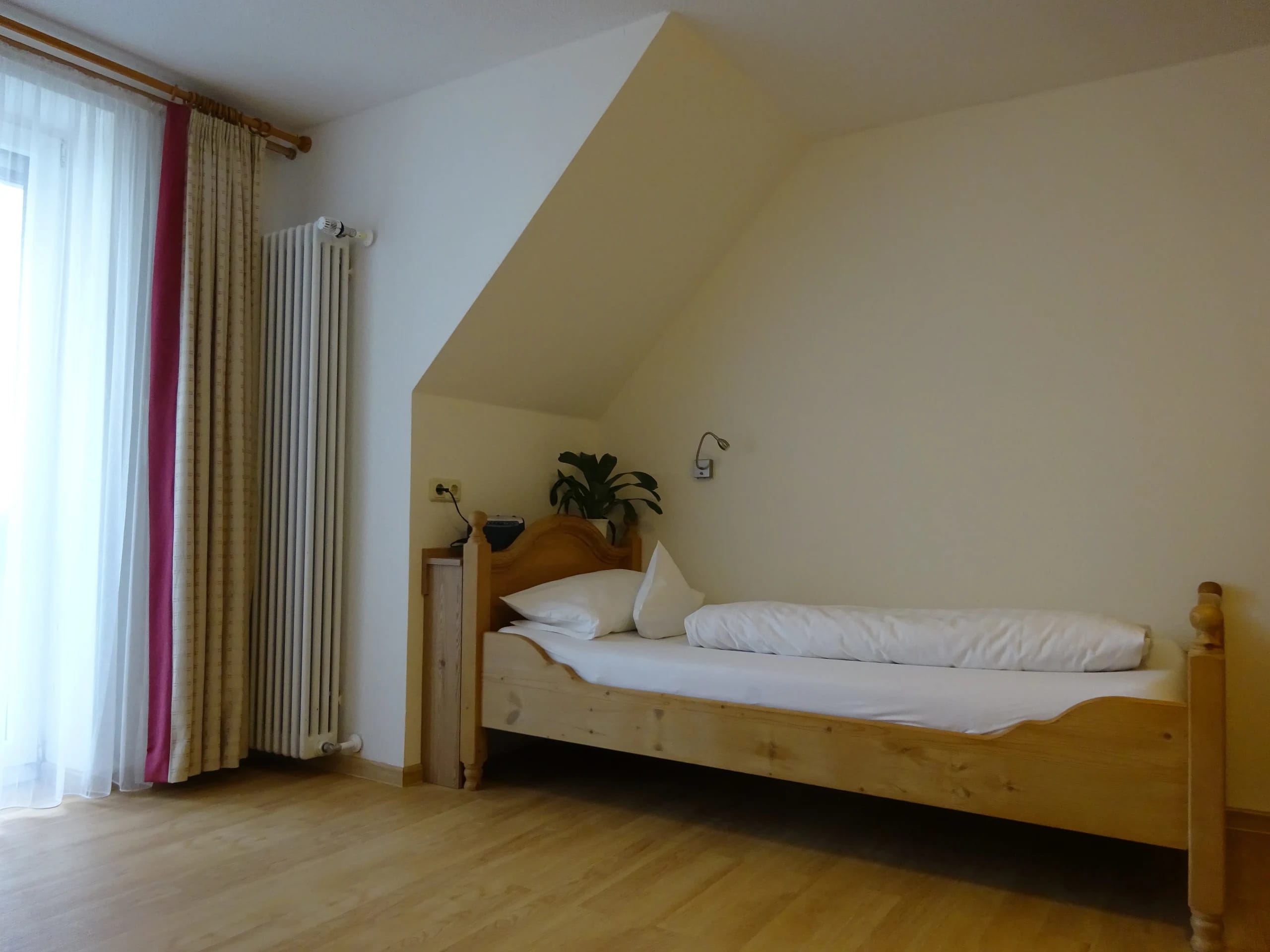 Einzelzimmer unter Dachschräge mit hellem Holzbett, Holzboden, Grünpflanze und bodenlangem Vorhang