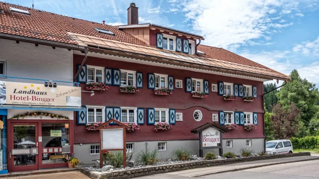 Straßenseitige Außenansicht des Hauses mit dunkelroter Fassade, blau-weiß verzierten Fensterläden, Blumenkästen und rotem Ziegeldach