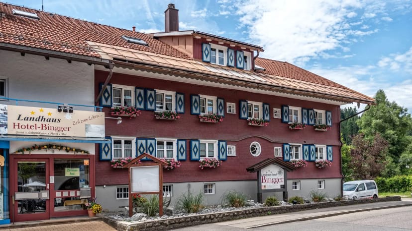 Straßenseitige Außenansicht des Hauses mit dunkelroter Fassade, blau-weiß verzierten Fensterläden, Blumenkästen und rotem Ziegeldach
