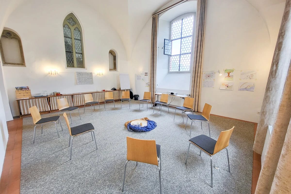 Heller Gruppenraum mit gotischen Fenstern, im Kreis gestellten Stühlen und einem Sitzkissen in der Mitte