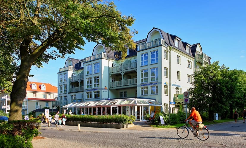 Klassische Fassade des Aparthotels mit Balkonen unter blauem Himmel, davor ein Baum und die Straße