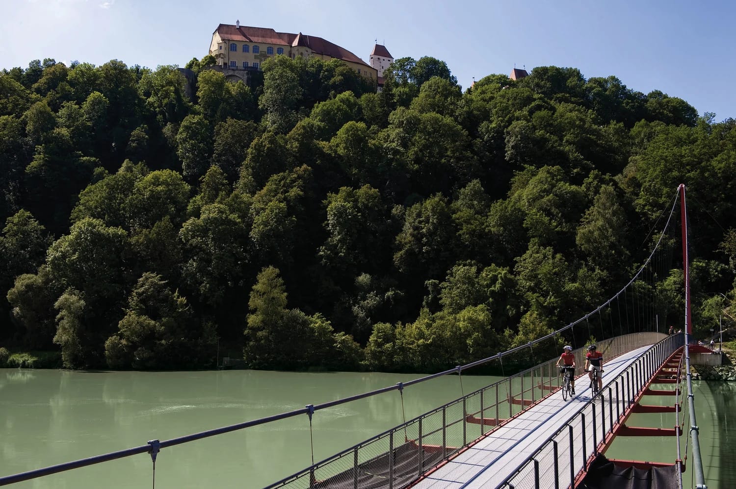 Hängebrücke über den Inn mit bewaldetem Ufer und Burg auf dem Hügel
