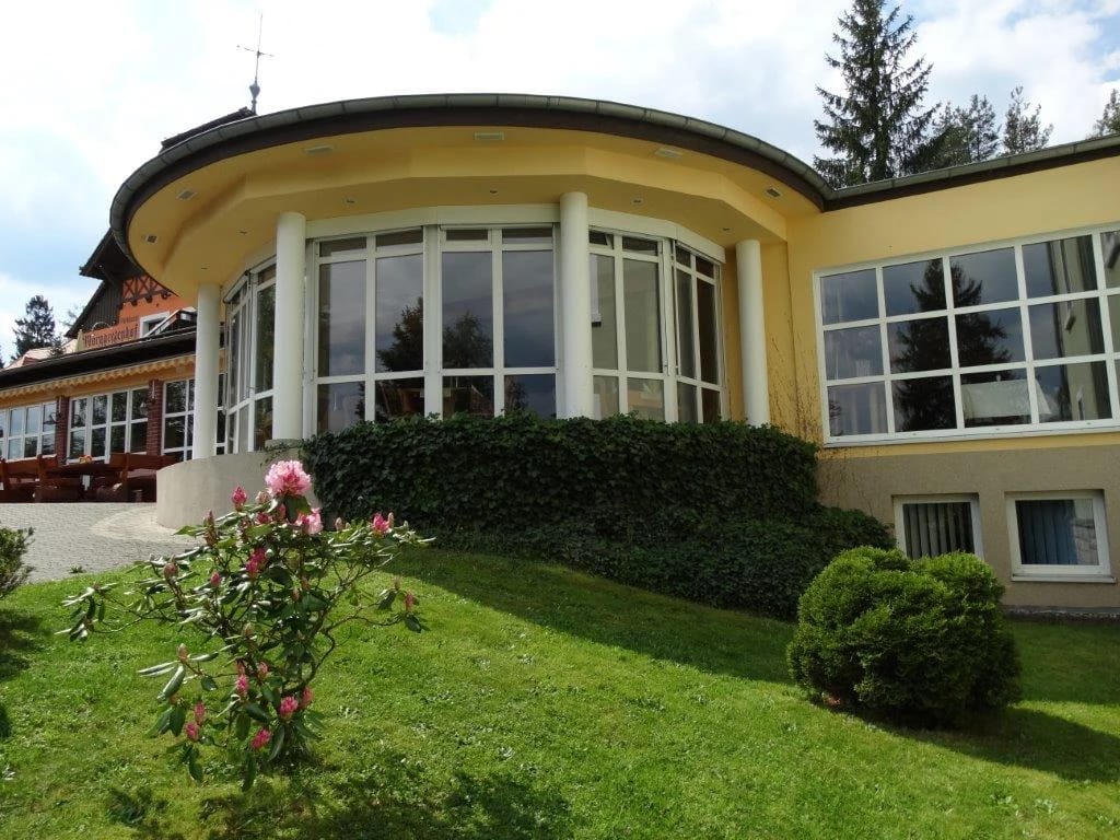 Runder Gebäudeteil mit umlaufender Fensterfront und geschwungenem Dach, davor blühender Rhododendron und Rasen