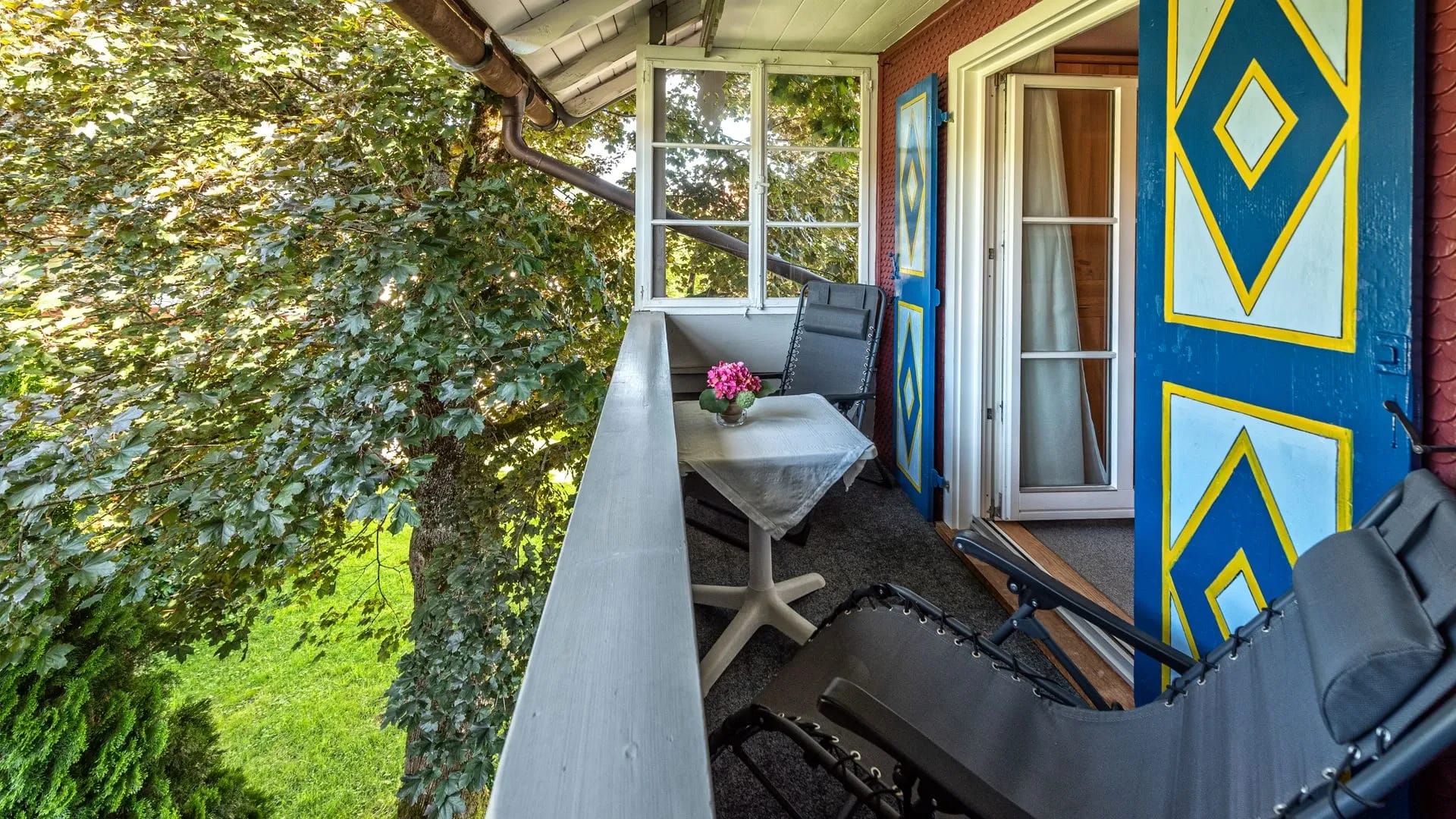 Balkon mit Liege, Tischchen mit Blumen und Stuhl, daneben die blau-gelb bemalte Hausfassade und Blick ins Grüne