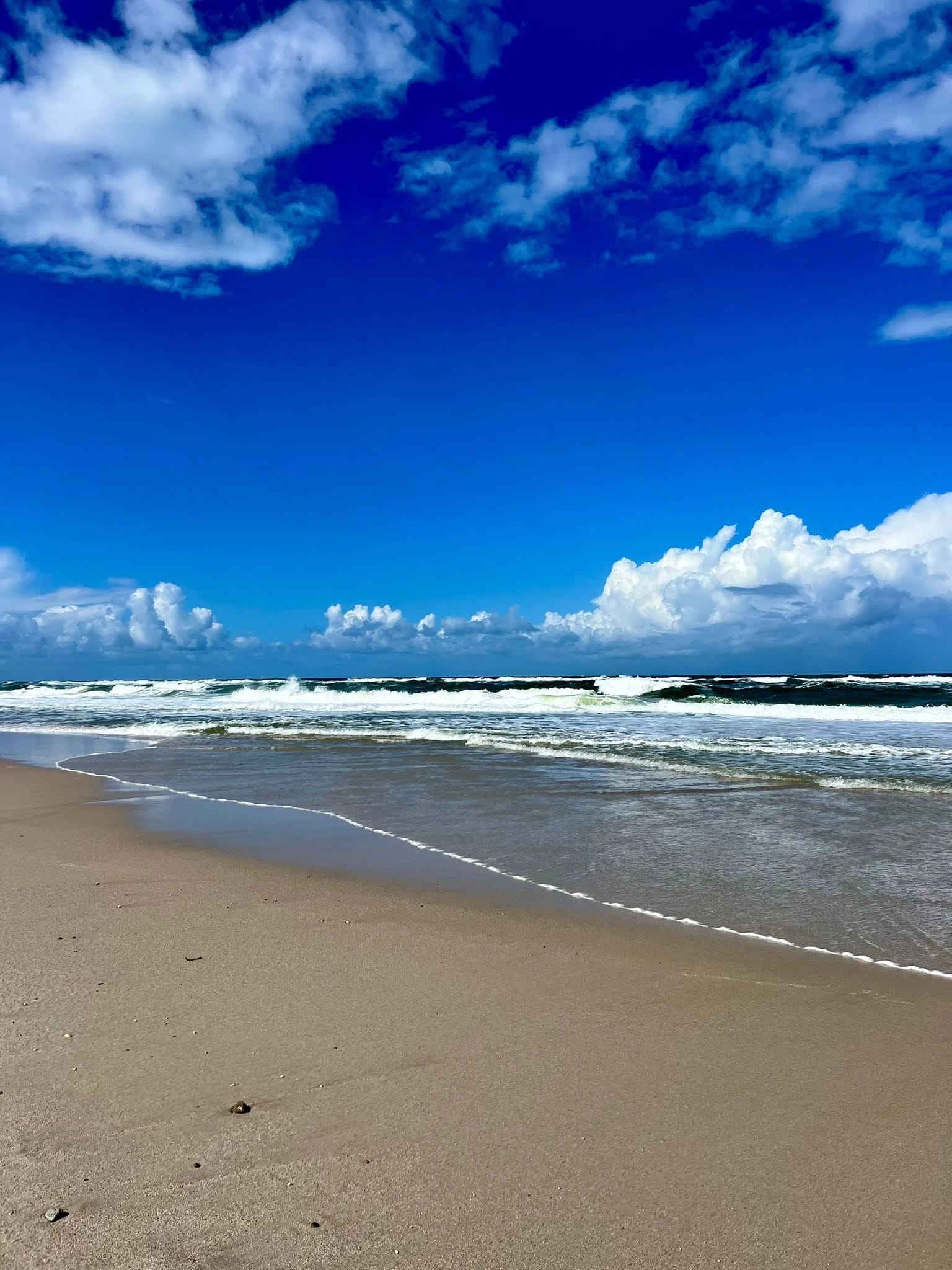 Breiter Sandstrand mit Brandung unter blauem Himmel mit Schönwetterwolken